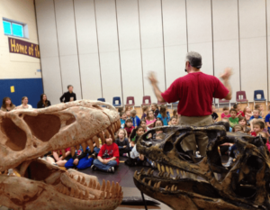 Dinosaurs Rock Instructor speaking to students at a Dinosaurs Rock School Assembly featuring 2 giant Dinosaur Skulls of a Allosaurus and a Tarbosaur.