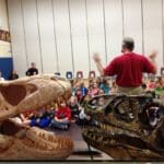 dinosaurs rock instructor speaking to students with a Tarbosaur Dinosaur Skull and a Allosaurus Dinosaur Skull featured in front